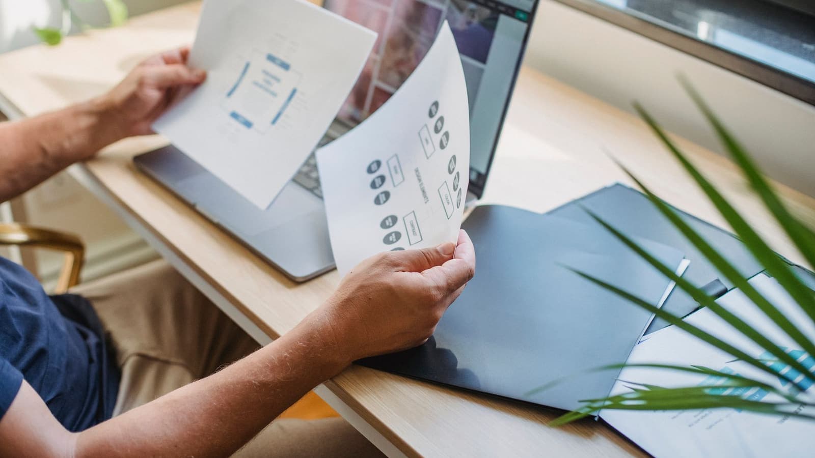 A designer comparing printed website layout sketches next to an open laptop.
