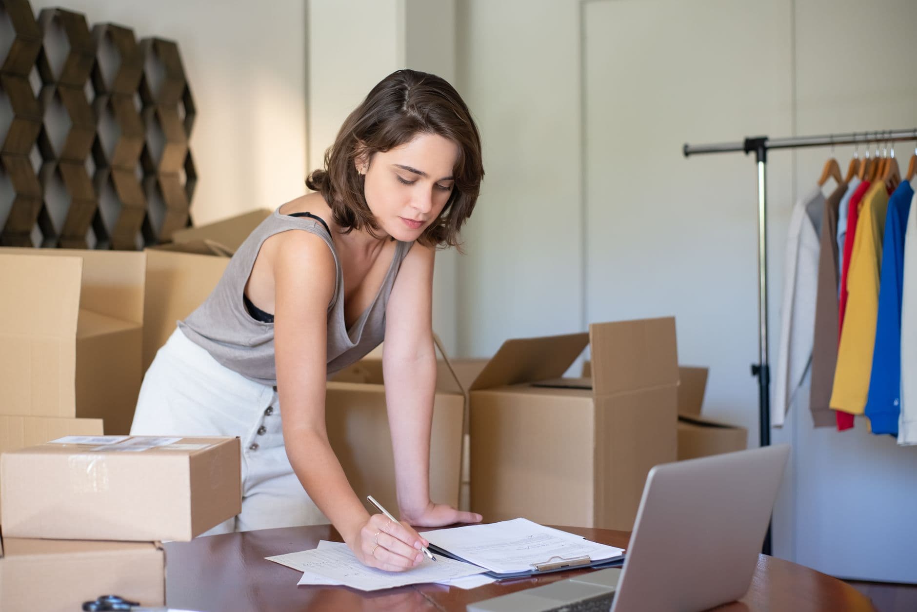 A small online store owner standing among shipping boxes, writing on an order slip with her laptop open on the desk.