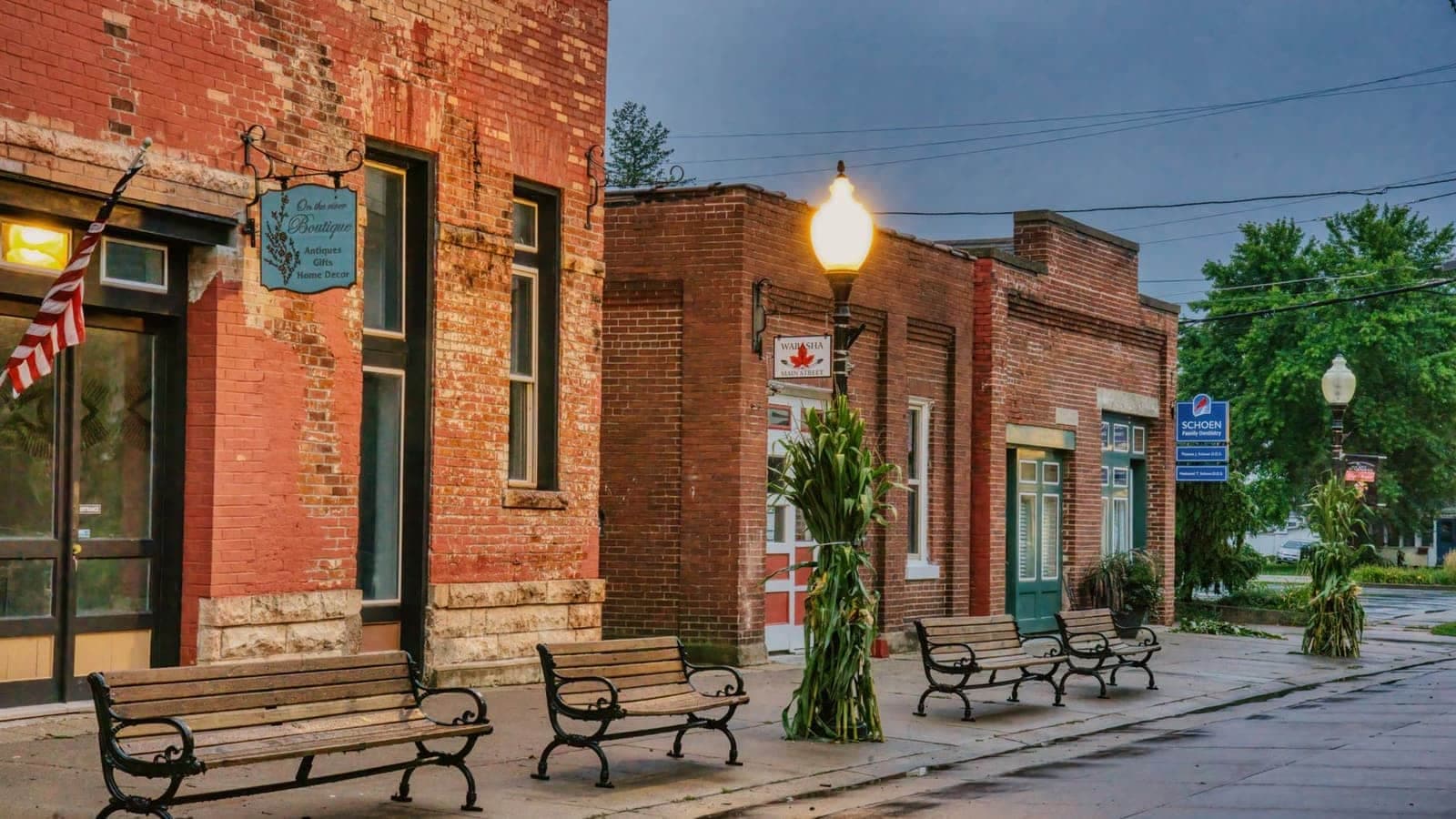 Brick storefronts, benches, and a flag along a small-town main street at dusk.