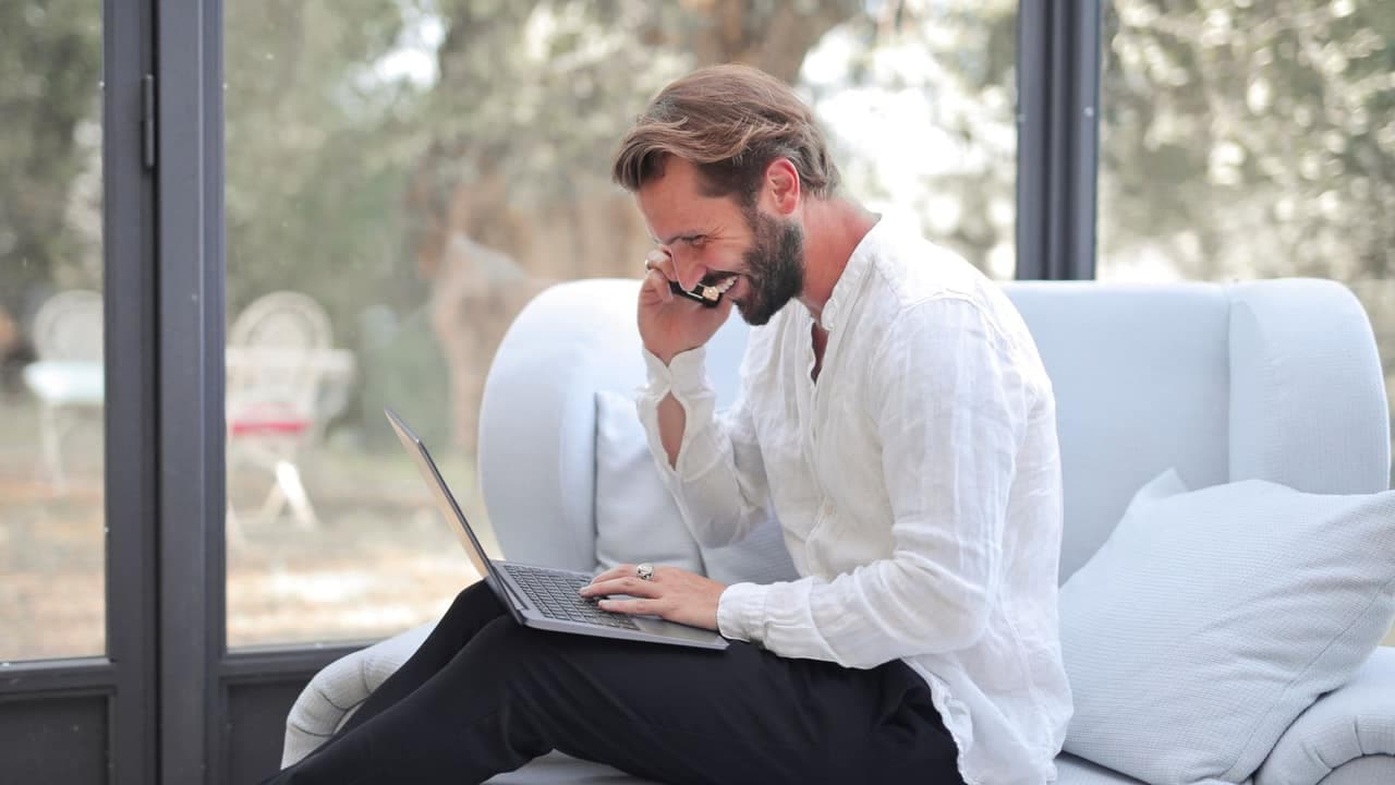 A man working from home, laughing on a phone call with a laptop on his lap